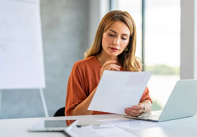 A women sitting in front of a laptop reading a document that she's holding.