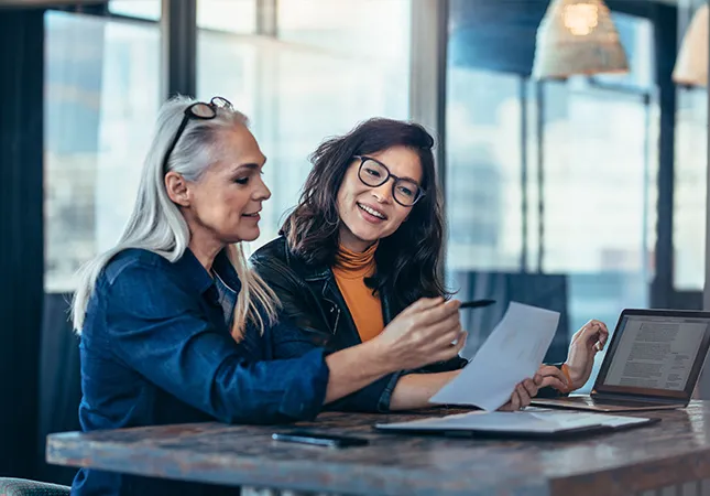 Two women sitting next to a laptop and discussing a document.