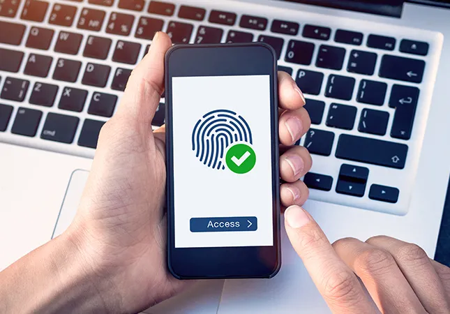 A person holding a phone over a laptop with a fingerprint icon on the screen of the phone.